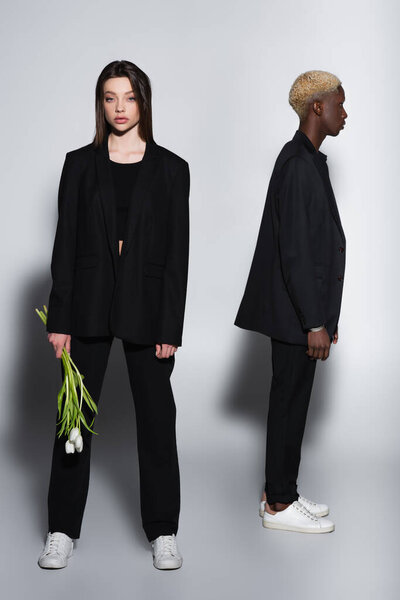 Брюнетка стильная женщина, стоящая с белыми тюльпанами рядом с блондинистым африканским мужчиной на сером с тенью