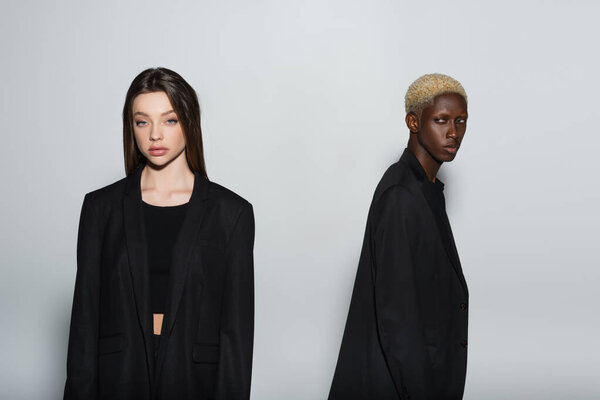 брюнетка женщина в черной одежде смотрит в камеру рядом с блондинкой африканский американец мужчина изолирован на сером
