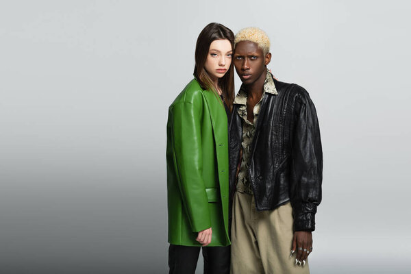межрасовая пара в модной одежде смотрит в камеру, стоя на сером