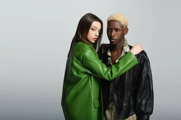 sensual woman in green leather jacket looking at camera and hugging blonde african american man isolated on grey