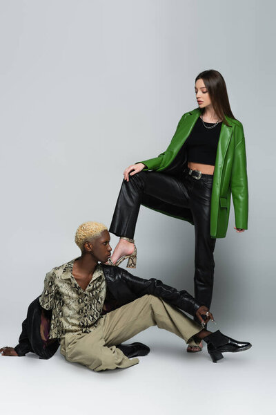 stylish brunette woman stepping on blonde african american man sitting on grey