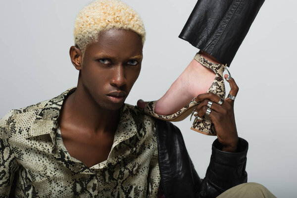 блондин африканский американец в футболке из змеиной кожи, смотрящий в камеру рядом с женщиной в сандалии, изолированной на сером