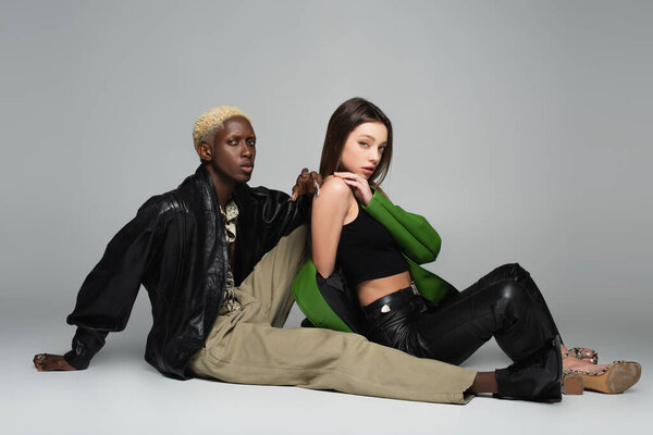 блондин африканский амейканец мужчина и чувственная брюнетка женщина сидит и смотрит в камеру на серый