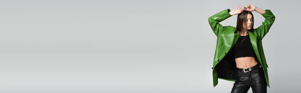 stylish woman in leather clothes posing with hands above head isolated on grey, banner