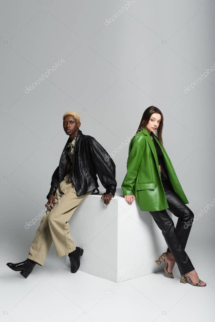 full length view of young and stylish multiethnic couple posing on white cube on grey