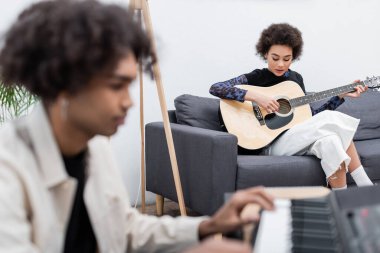 Evde sentezleyici ile bulanık erkek arkadaşının yanında akustik gitar çalan Afrikalı Amerikalı bir kadın. 