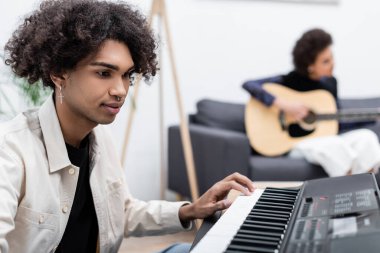 Young african american man playing synthesizer near blurred girlfriend with acoustic guitar at home  clipart