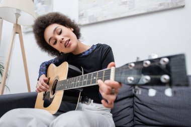Genç Afrikalı Amerikalı kadın oturma odasındaki kanepede akustik gitar çalıyor. 