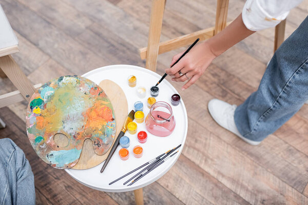 Cropped view of african american woman holding paintbrush near paints and easels at home 