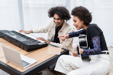 Gülümseyen Afro-Amerikan kadın akustik gitarıyla erkek arkadaşını işaret ederek evde sentezleyici ve dizüstü bilgisayar çalıyor. 