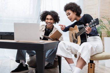 Gülümseyen Afro-Amerikan adam evde akustik gitarlı, sentezleyici ve kız arkadaşının yanındaki bulanık laptopa bakıyor. 