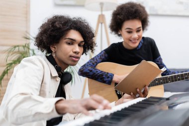 Gülümseyen Afro-Amerikan kadın akustik gitar çalıyor. Yanında sentezleyici ve not defteriyle sevgilisinin yanında. 