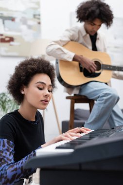 Genç Afrikalı Amerikalı müzisyen evde akustik gitarlı bulanık erkek arkadaşının yanında sentezleyici çalıyor. 