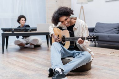 Neşeli Afrikalı Amerikalı adam evde bulanık kız arkadaşının yanında akustik gitar çalıyor. 