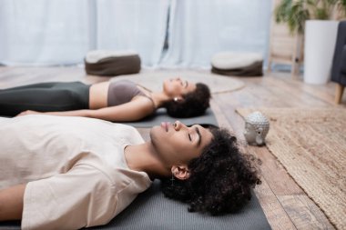 Genç Afrikalı Amerikalı adam evde yoga minderinde bulanık kız arkadaşının yanında meditasyon yapıyor. 