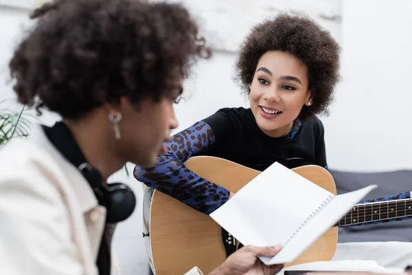 Lächelnde Afrikanerin Spielt Akustikgitarre Neben Verschwommenem Freund Mit Notizbuch Hause — Stockfoto