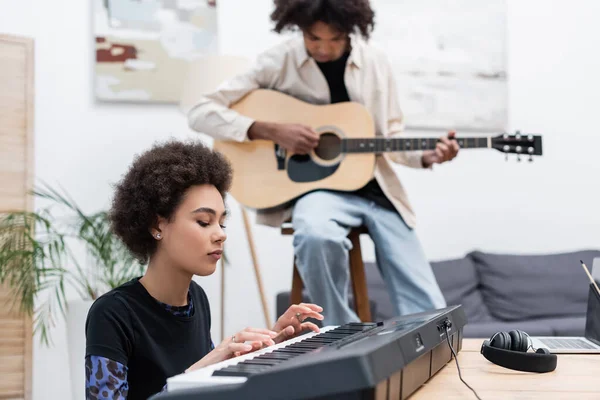 在笔记本电脑附近玩合成器的非洲裔美国女人 在家里带着声吉他的模糊男友 — 图库照片