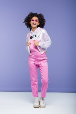 Pembe güneş gözlüklü ve tulumlu mutlu Afro-Amerikalı kadın mor üzerine moda ceketi ayarlıyor.