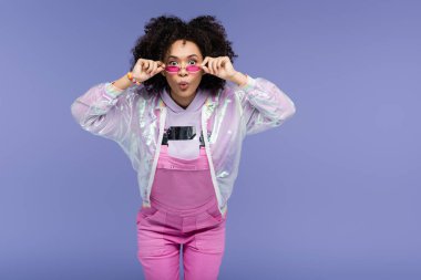 surprised african american young woman in pink overalls adjusting sunglasses isolated on purple clipart