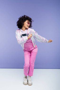 Uzun boylu, gülümseyen Afro-Amerikan kadın. Mor tulumlar içinde poz veriyor.