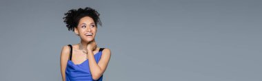 cheerful african american woman in bright blue dress looking away while posing isolated on grey, banner clipart