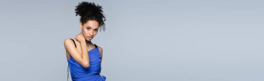 Kıvırcık Afro-Amerikan modeli, parlak mavi elbisesiyle kameraya bakarken gri pankartta yalnız poz veriyor.