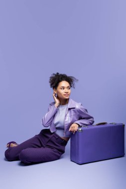 Uzun boylu, rüya gibi Afro-Amerikan modeli deri ceketli menekşe rengi bavulun yanında mor üzerinde oturuyor. 
