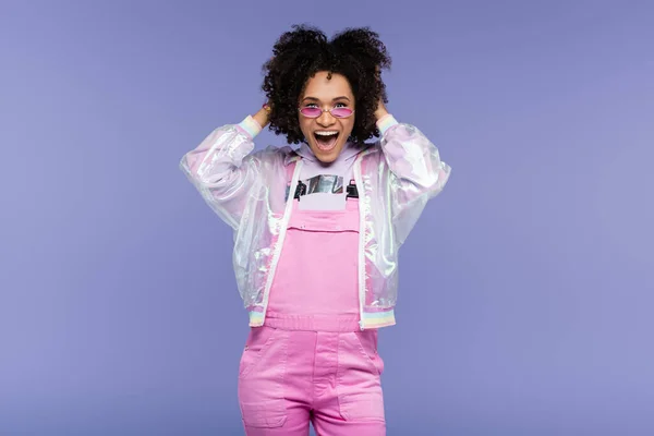 excited african american woman in stylish sunglasses and overalls adjusting curly hair isolated on purple