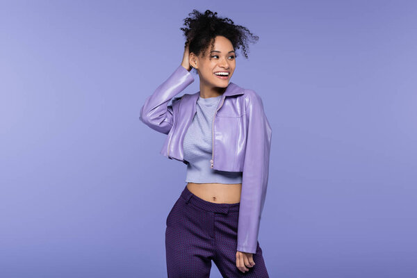 trendy african american woman in leather jacket smiling and looking away isolated on purple