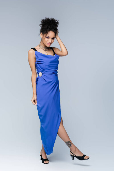 full length of smiling and tattooed african american woman in bright blue dress on grey