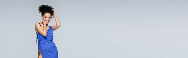 довольная молодая африканская американка в ярко-синем платье позирует изолированно на сером, баннер