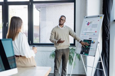 Gözlüklü Afrikalı Amerikalı iş adamı, kağıt bardakla meslektaşlarına çizelgeleri gösteriyor.