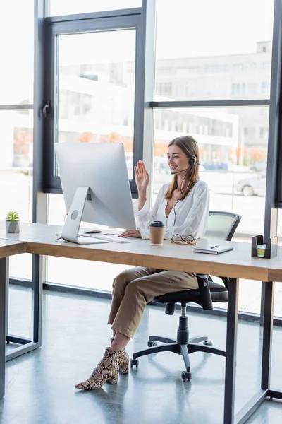 happy woman in headset waving hand and looking at computer monitor in office