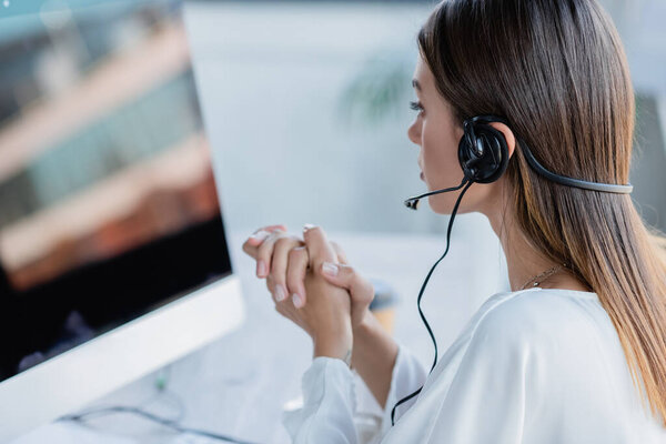 young businesswoman in headset sitting with clenched hands and looking at monitor 
