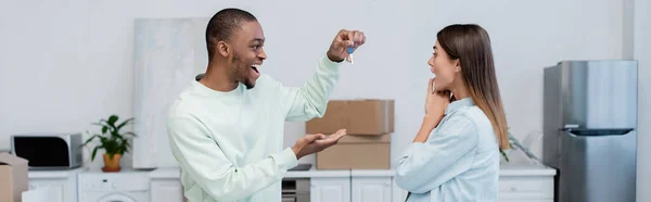 新しいアパートで興奮したガールフレンドの近くに鍵を持っている幸せなアフリカ系アメリカ人男性バナー — ストック写真