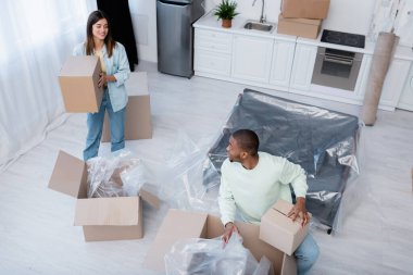 high angle view of pleased interracial couple unpacking boxes during relocation  clipart