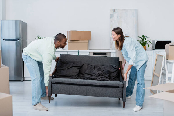 happy interracial couple carrying sofa in living room 