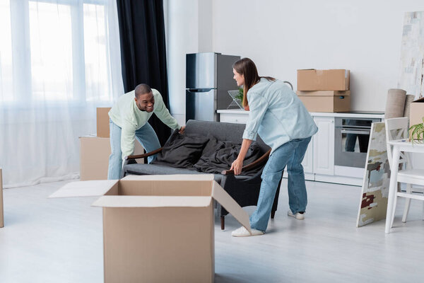 cheerful interracial couple moving sofa in living room 