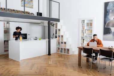 Neşeli Afrikalı Amerikalı kadın erkek arkadaşı evde çalışırken sandviç yapıyor. 