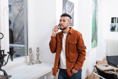 curly african american man talking on cellphone while standing with hand in pocket in modern apartment  clipart