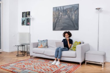 Genç Afrikalı Amerikalı kadın koltukta oturuyor ve dizüstü bilgisayara bakarken cep telefonundan sesli mesaj kaydediyor. 