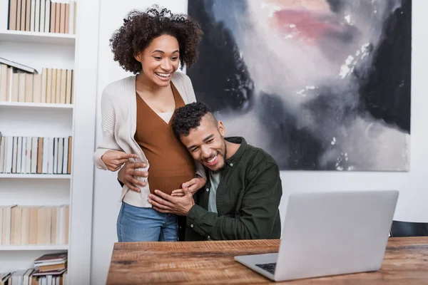 ラップトップでのビデオ通話中に幸せな夫の近くで腹を指している陽気な妊娠中のアフリカ系アメリカ人女性 — ストック写真