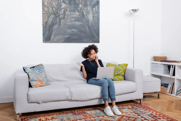 Joven Afroamericana Mujer Sentada Sofá Hablando Teléfono Inteligente Cerca Computadora — Foto de Stock