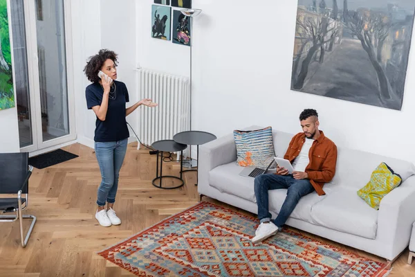 若いアフリカ系アメリカ人の女性が携帯電話で話している間にボーイフレンドは現代のアパートでデジタルタブレットを使用して — ストック写真