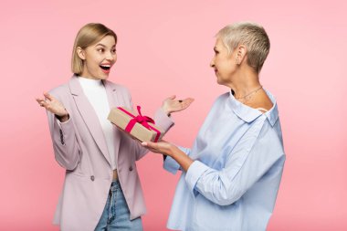 mature mother giving present to amazed daughter isolated on pink clipart