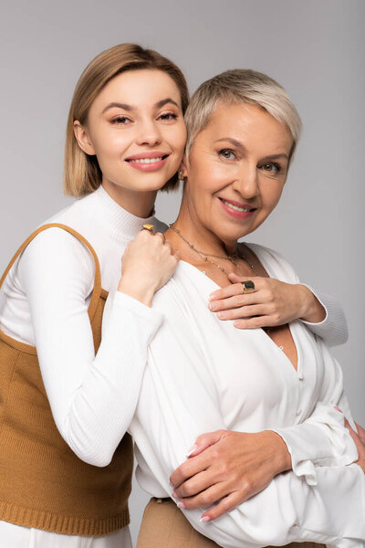 happy young daughter hugging middle aged mother and looking at camera isolated on grey