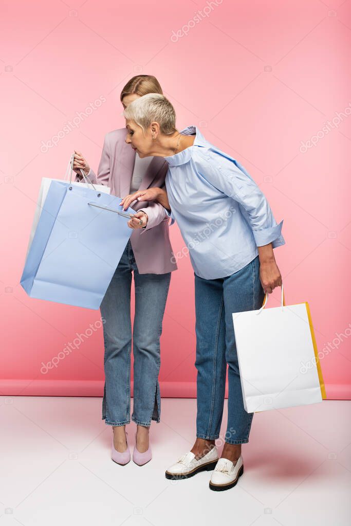 full length of young woman and mature mother in jeans looking at shopping bags on pink