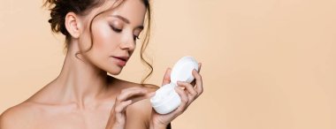 Young model with naked shoulders holding cosmetic cream and cap isolated on beige, banner  clipart