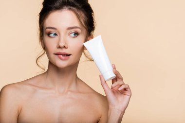 Bej üzerine izole edilmiş kozmetik losyon tutan çıplak omuzlu düşünceli bir model. 
