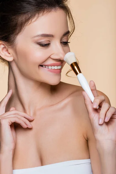 Happy woman with naked shoulder holding cosmetic brush near nose isolated on beige